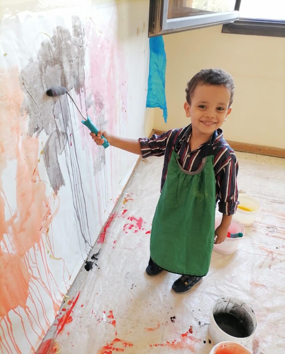 A young boy in a green smock smiles as he rolls paint onto a wall at Rifka's House.