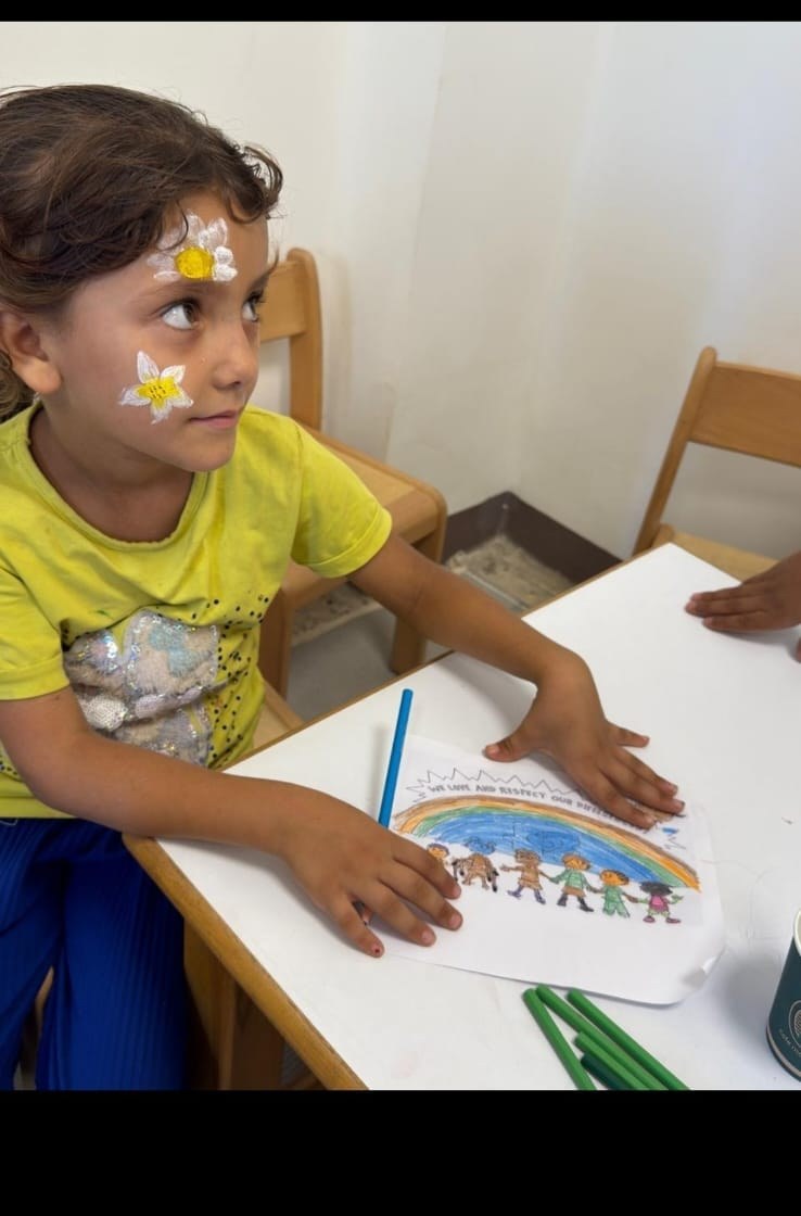 A young girl with painted flowers on her cheeks colors a rainbow drawing at Rifka's House.